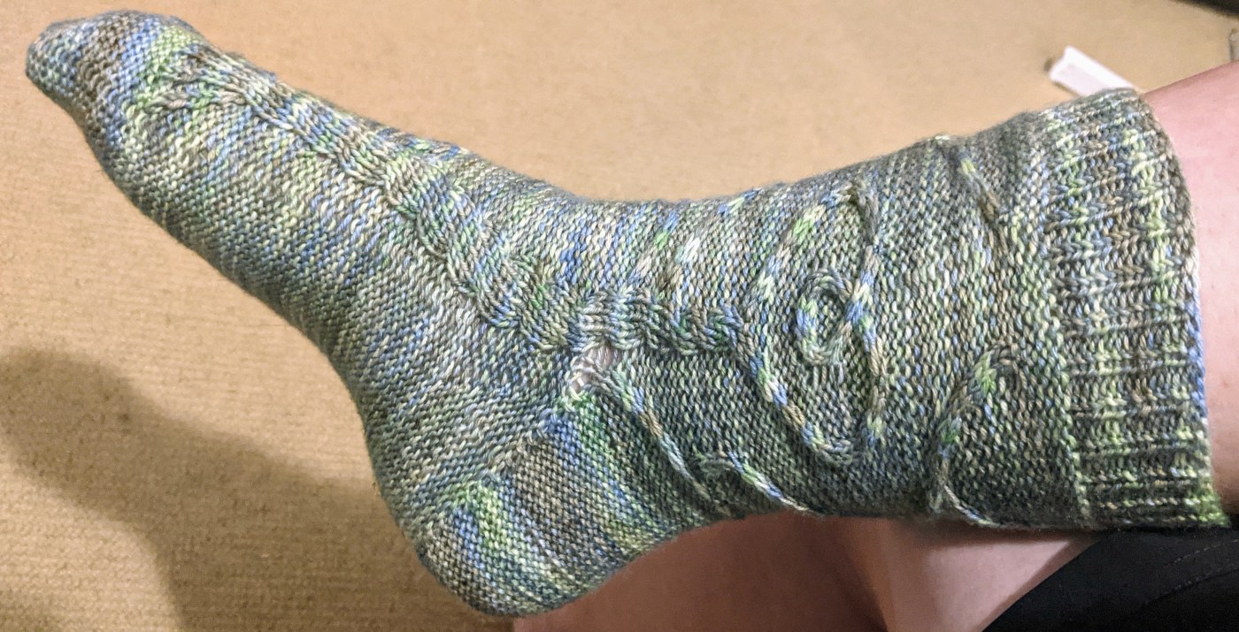 handknit sock on an adult foot. The pattern in the sock is a cabled design featuring a heart shape and a twisted tree like cable down the entire length from the heart.