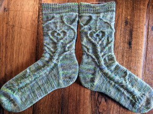 Hand knit socks depicting a mirrored tree design using cable stitches with a heart and branch detail. The yarn is earthy tones and light coloured overall