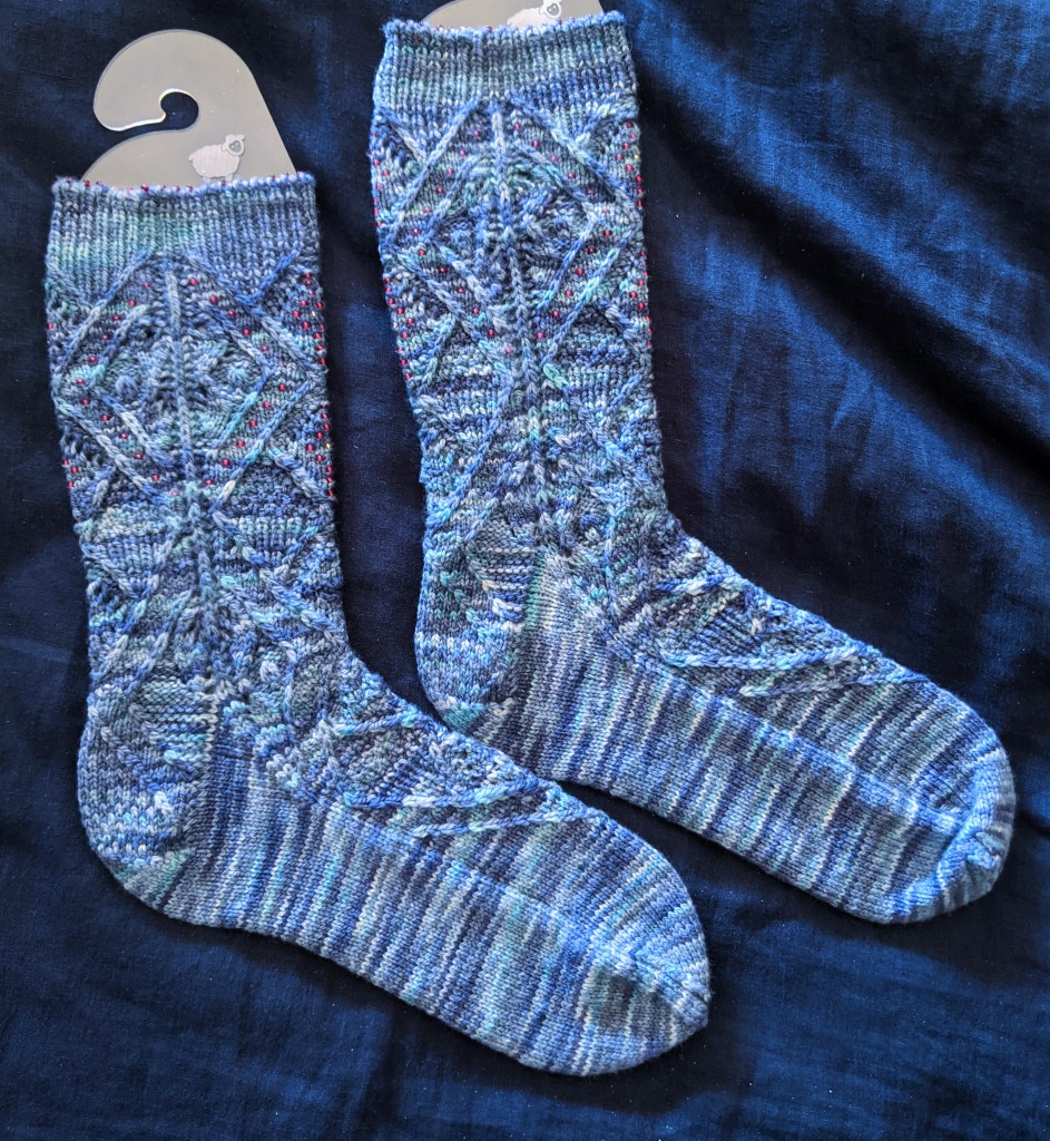 pair of hand knit socks viewed from the side. There is a diagonal lines pattern running the whole length that includes beads. The underside of the feet is plain stocking stitch.