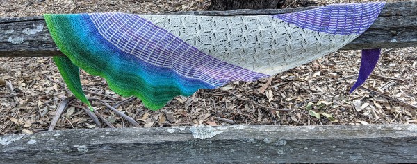 multi-yarn crescent shawl is draped over a railing made from old railway sleepers. The yarns are white and a gradient going from purple to green. There is a central panel of lace knit with the white yarn and a slip stitch design on either side of it using both yarns. Outside of that is just the gradient yarn for each end.